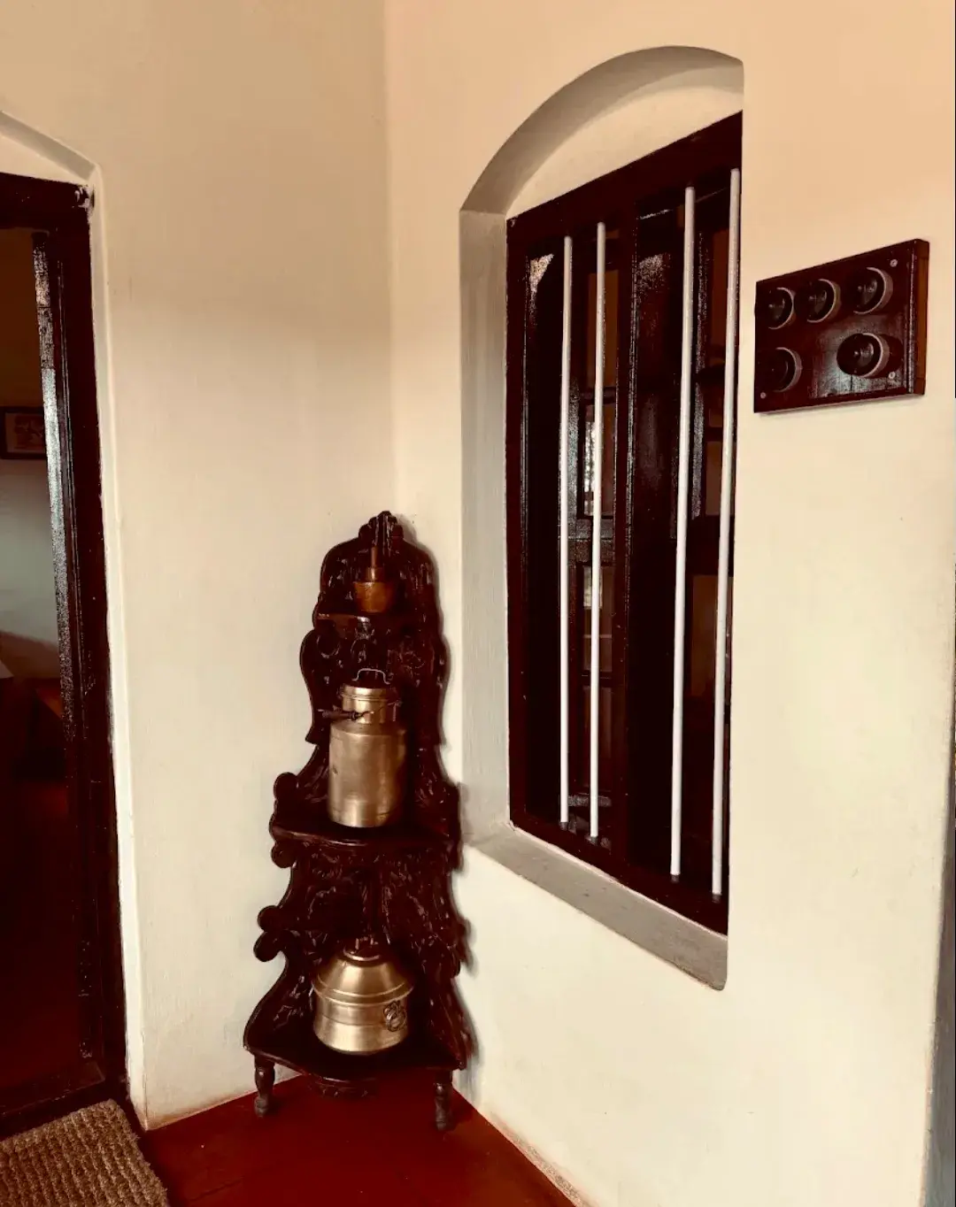 An arched window with dark wooden shutters and brass vessels on a carved stand.